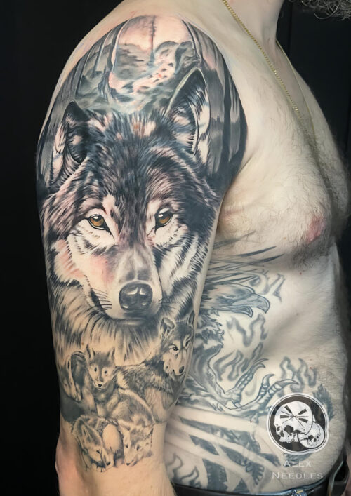 Tatouage d'une tête de loup réaliste avec une louve et ses petits en dessous et un décor de forêt sur le dessus. Réalisé sur un bras par Alex, The Needles Factory
