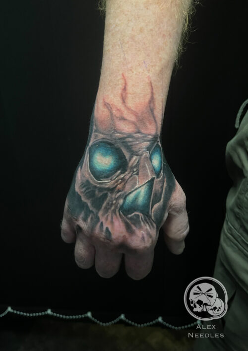 Crane Tatouage représentant un crane réalisant en noir et gris avec des touches de bleu dans les yeux et le nez, sur une main par Alex, The Needles Factory
