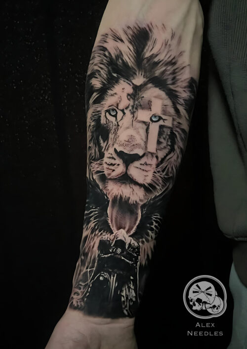 Tatouage représentant un lion au yeux bleu réaliste avec une croix au niveaux de l'oeil droit, avec un ange en noir sous la tête réalisé sur un avant bras par Alex, The Needles Factory